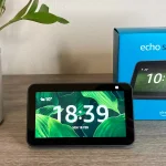 Un dispositivo Echo Show 5 posizionato su un mobile in salotto con sullo sfondo la sua scatola e una pianta decorativa