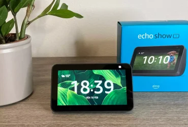 Un dispositivo Echo Show 5 posizionato su un mobile in salotto con sullo sfondo la sua scatola e una pianta decorativa